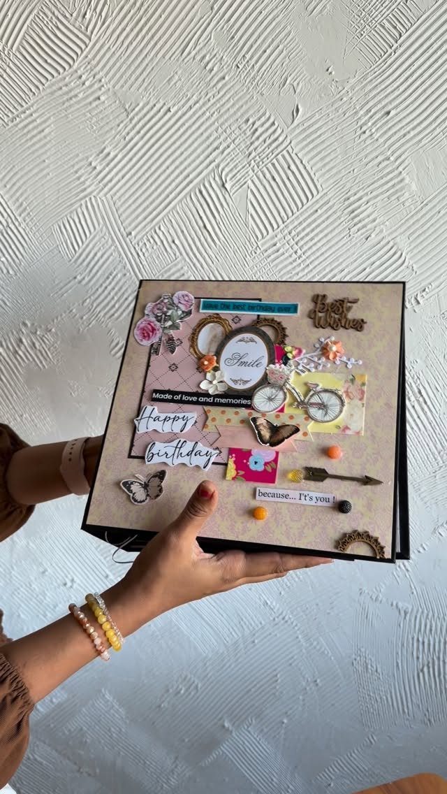 Hands holding a decorated handmade scrapbook with floral and butterfly embellishments against a white wall.