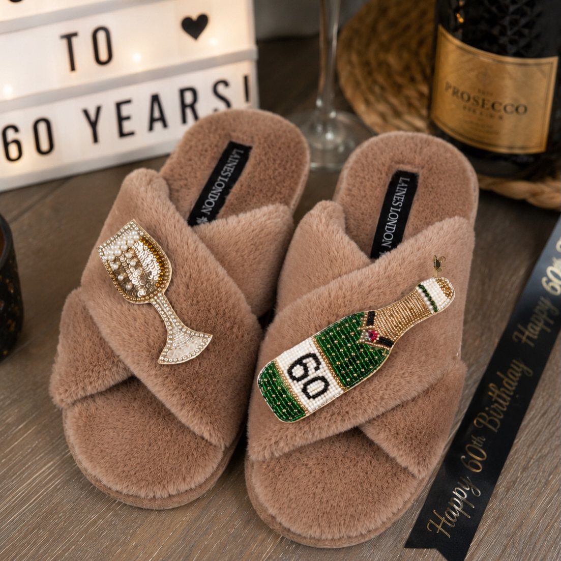 Tan fuzzy slippers decorated with rhinestone champagne flute and bottle embellishments on a wooden surface.