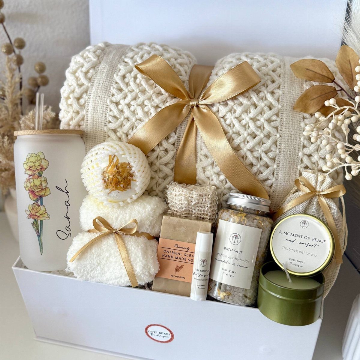 A white gift box containing a cream blanket, rolled towels, bath salts, and spa accessories.