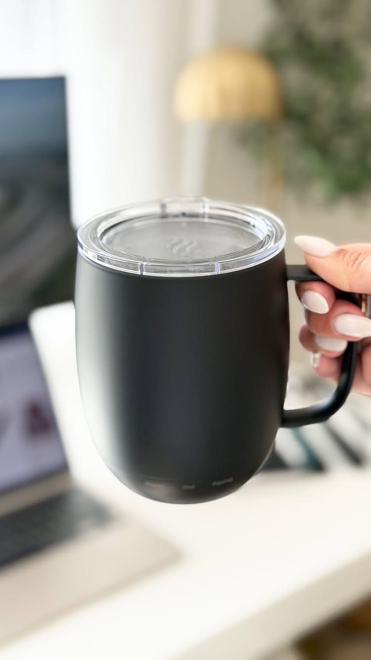 A hand holding a matte black smart mug with a clear lid in an office setting.