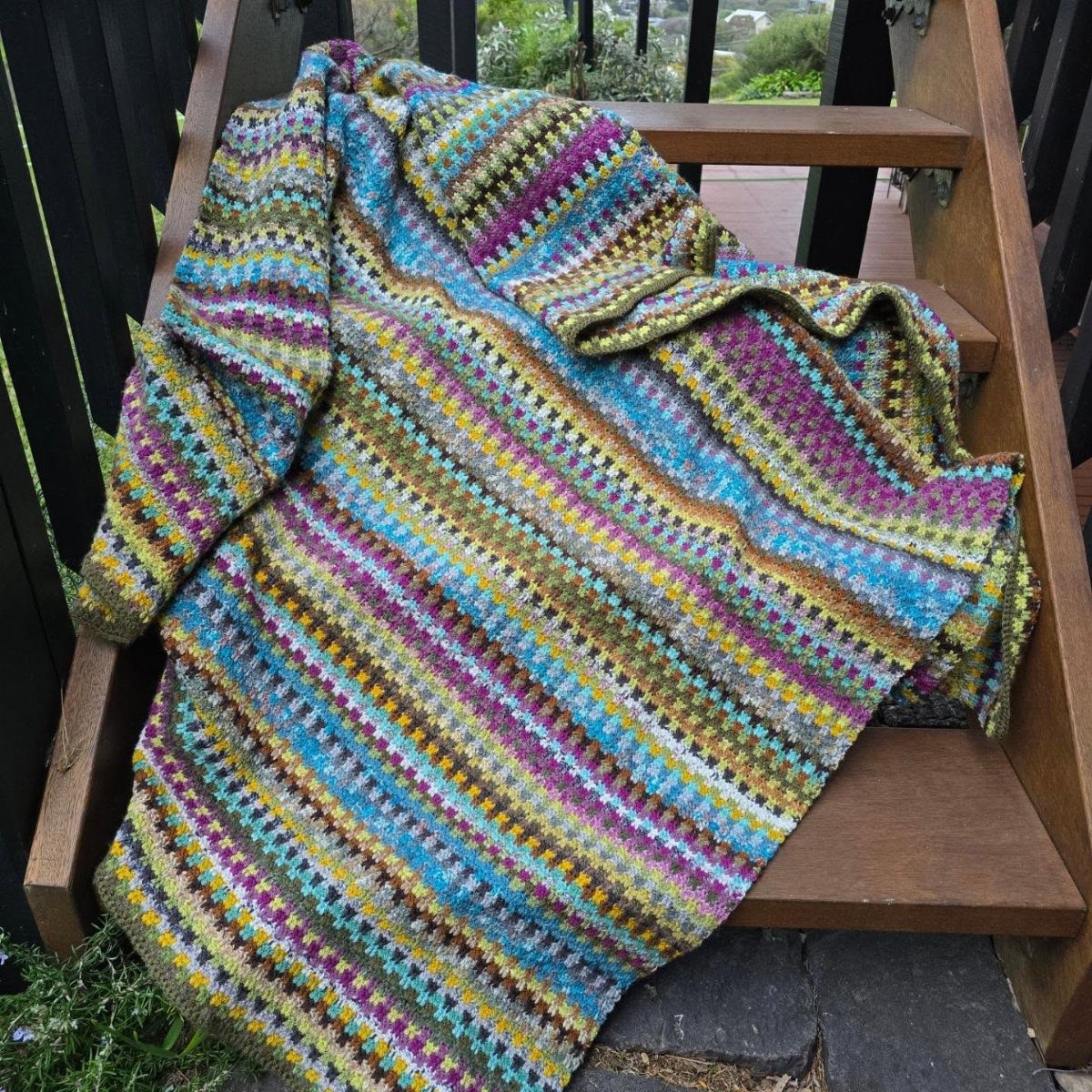 A colorful striped crocheted blanket draped over a wooden outdoor bench.