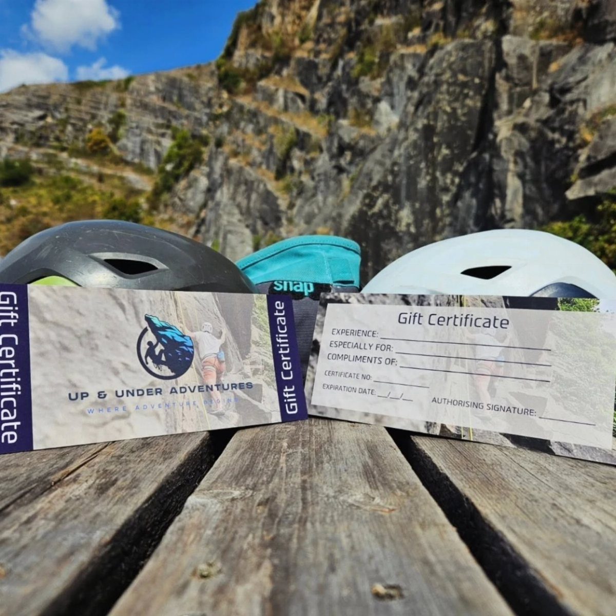 Two adventure gift certificates resting on a wooden table with climbing helmets and cliffs behind.