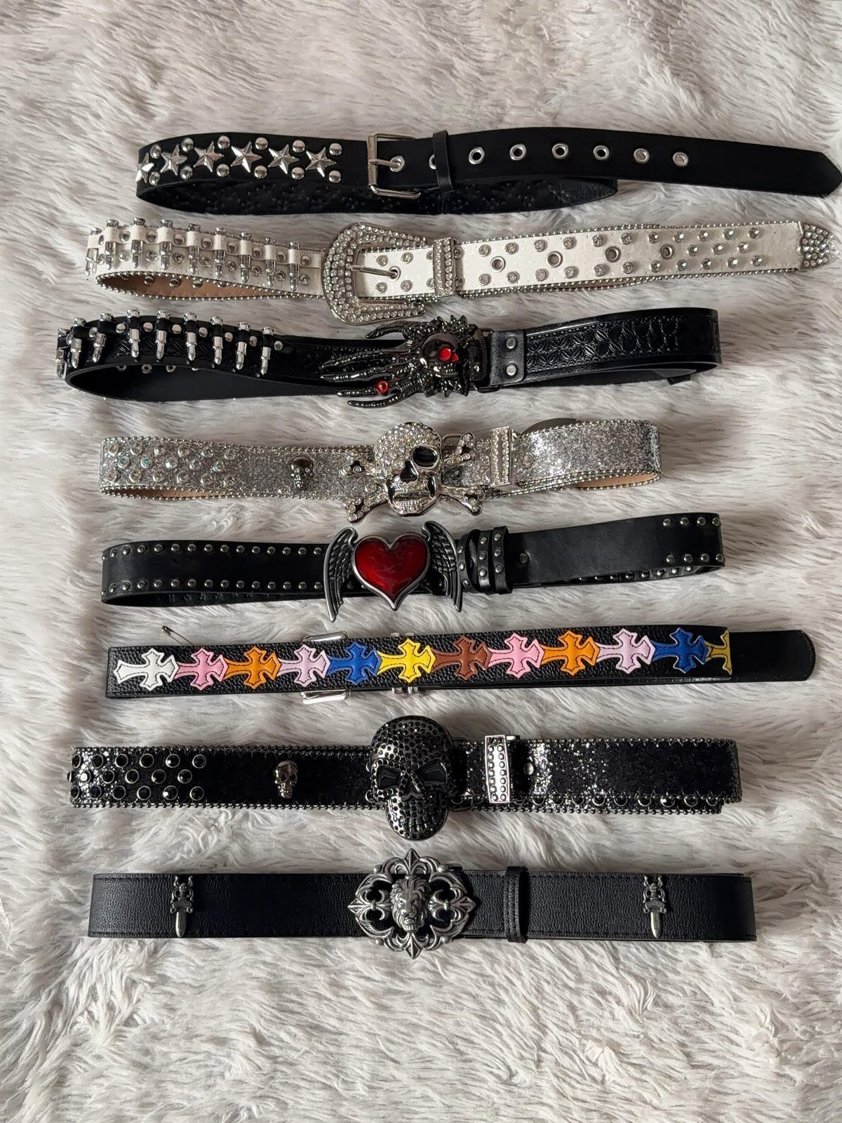Various decorative belts with studs, rhinestones, and skull buckles laid on white faux fur.
