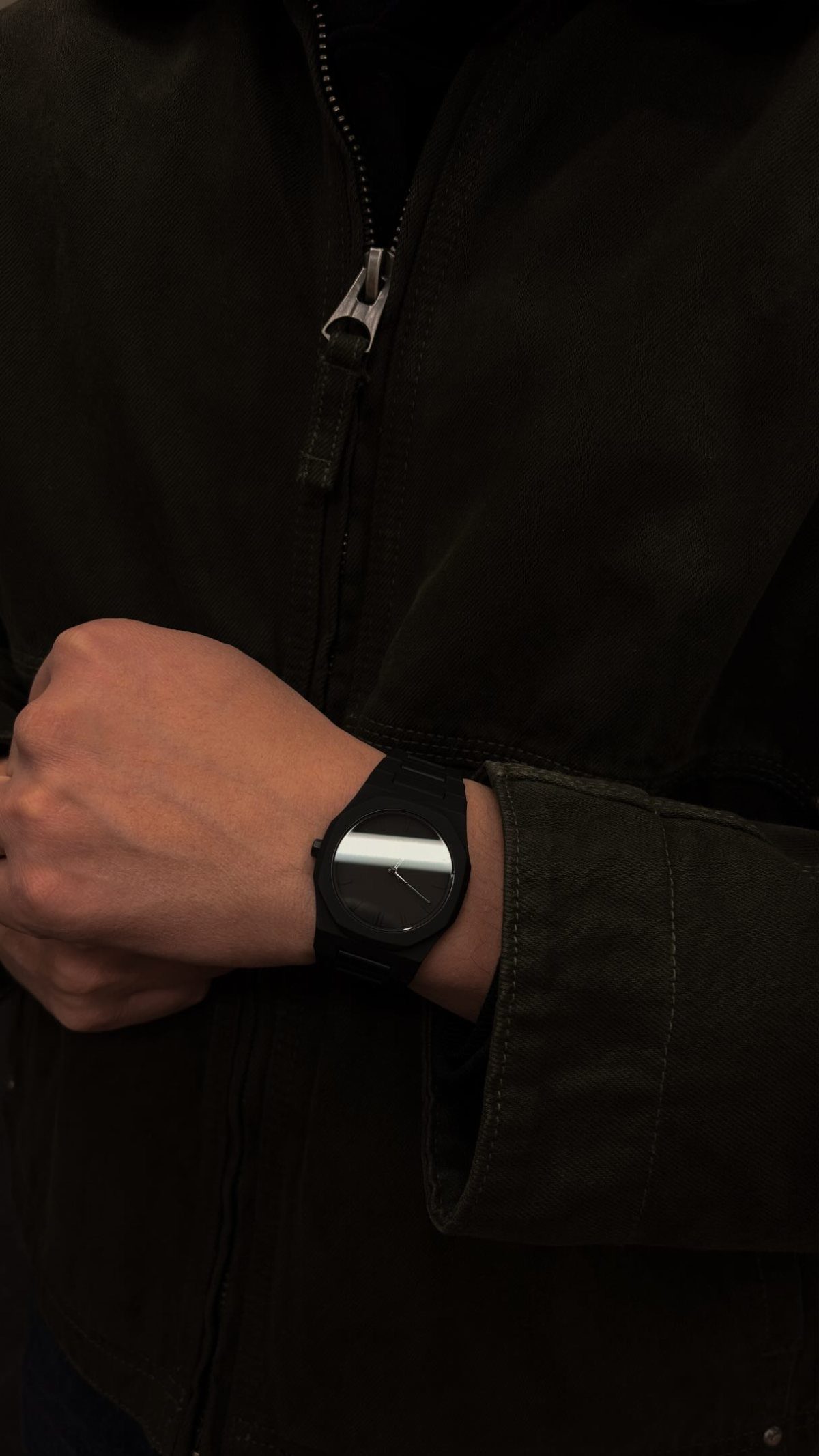 A person wearing a black wristwatch with a silver rectangular face and a dark jacket.