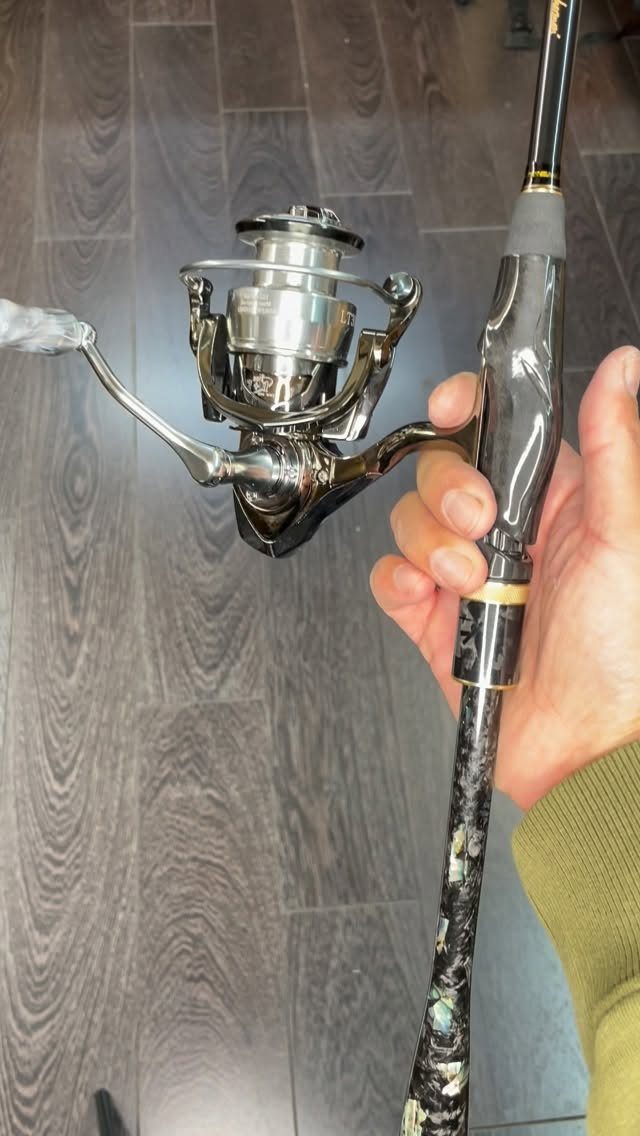A hand holding a black fishing rod with a silver spinning reel against a dark floor.
