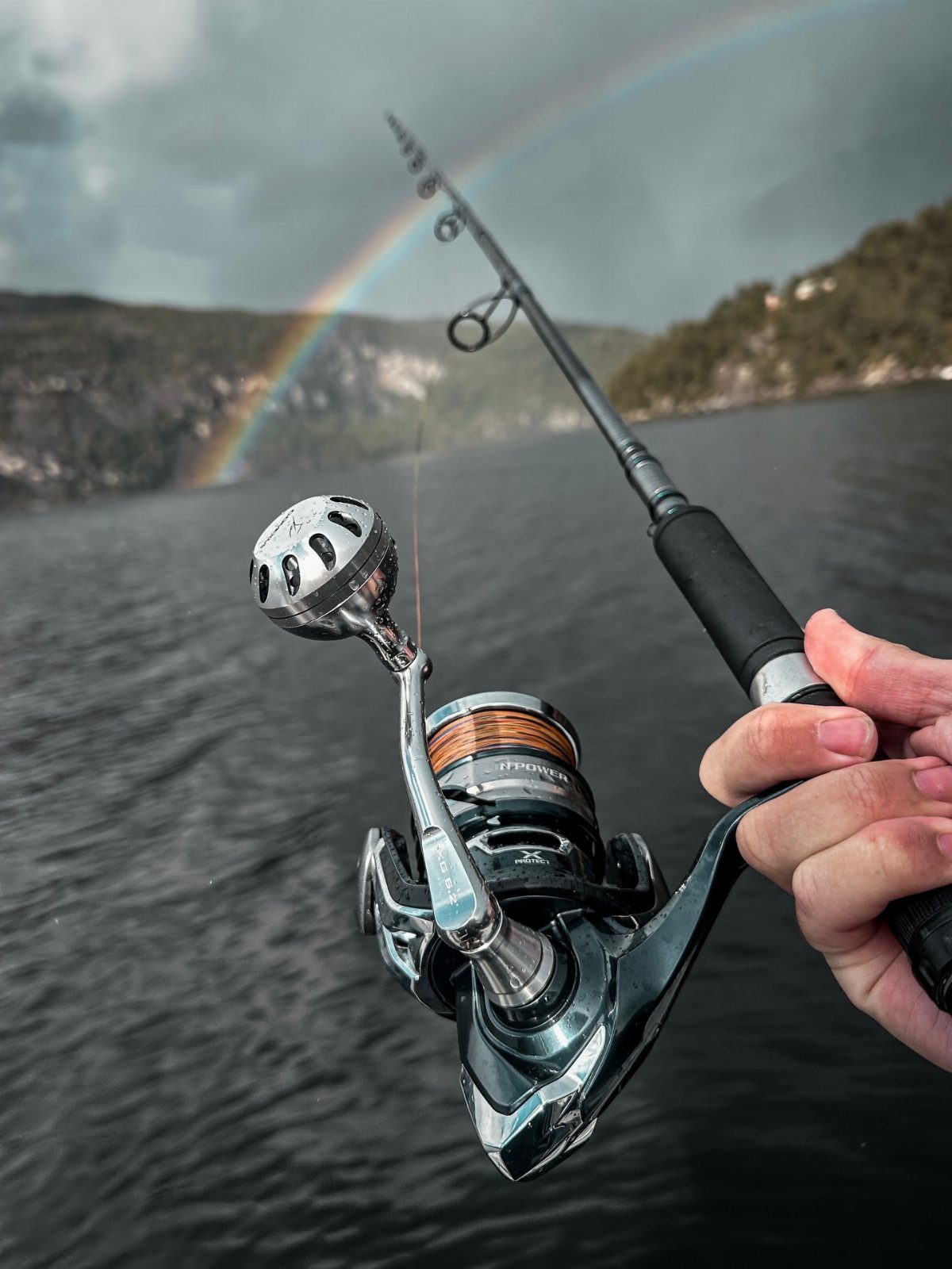 A hand holding a fishing rod and reel over a lake with a rainbow in the background.