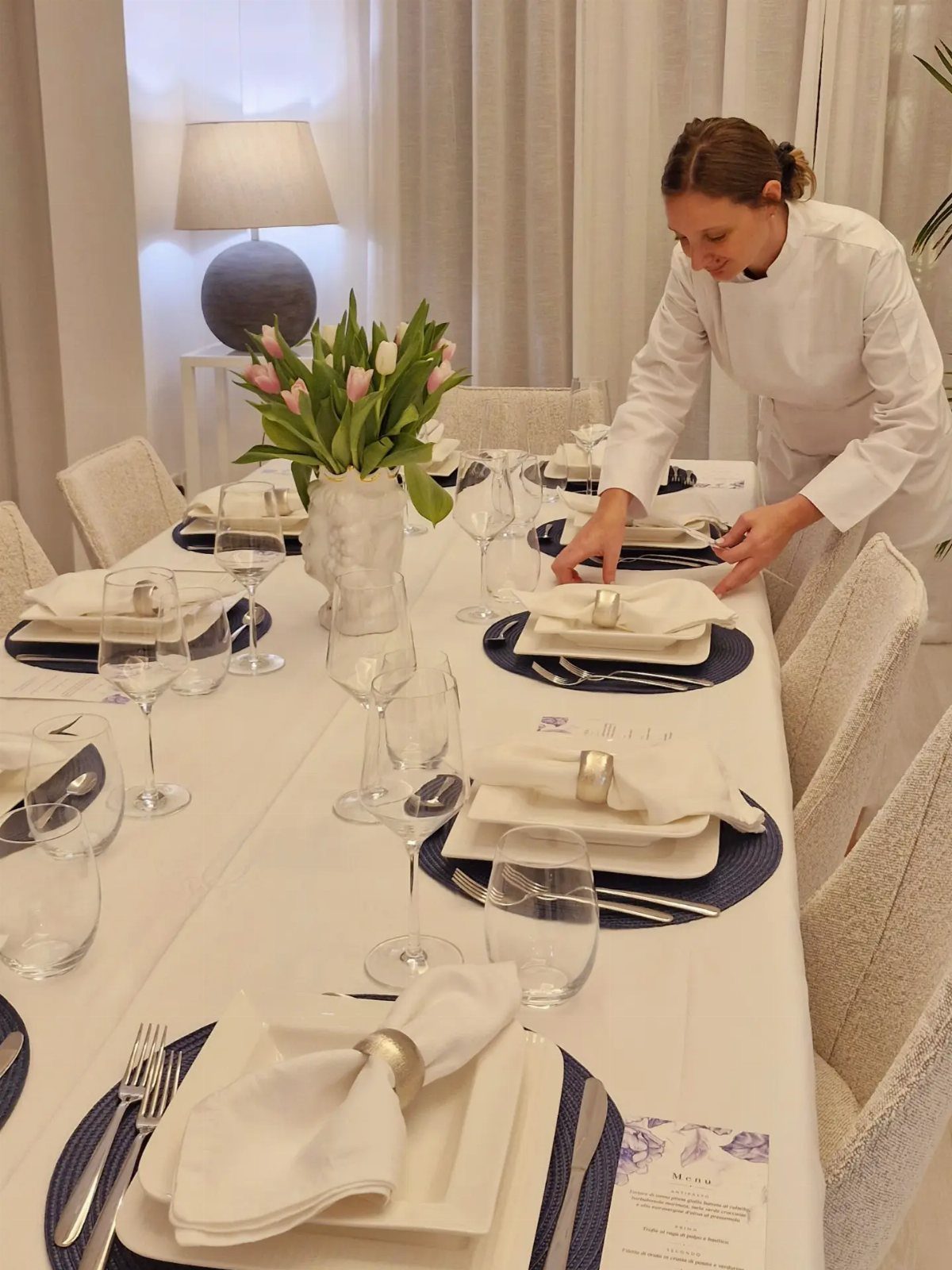 A chef in white clothing setting a formal dining table with elegant place settings and pink tulips.