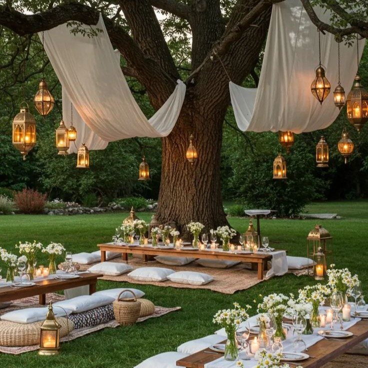 Low wooden dining tables with white flowers and candles set under a tree with lanterns.