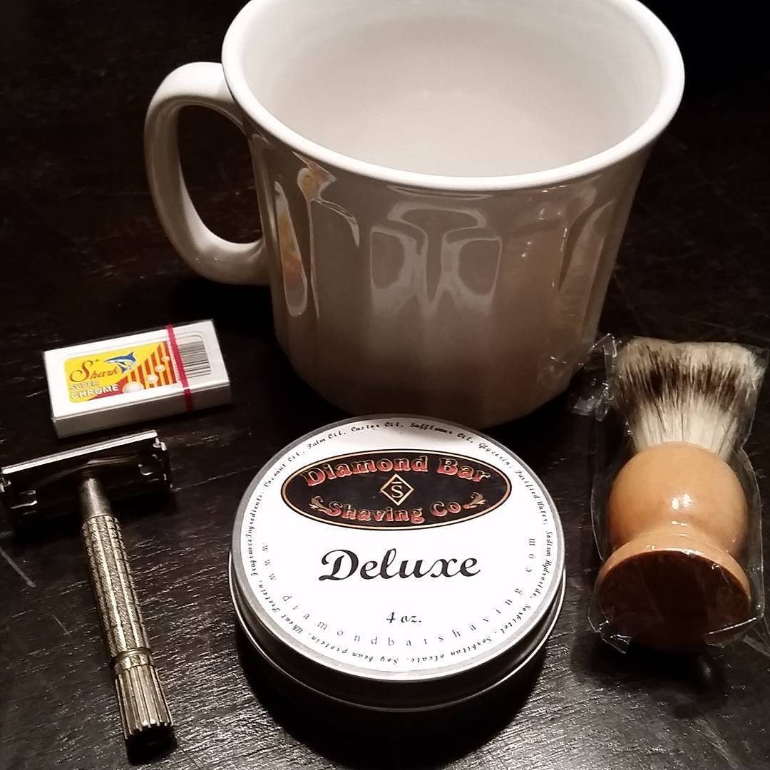 A safety razor, shaving brush, soap tin, and white mug arranged on a dark surface.
