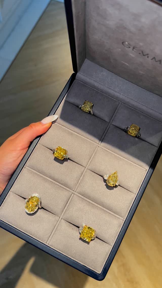 A hand holding an open grey velvet jewelry box containing six yellow gemstone rings.