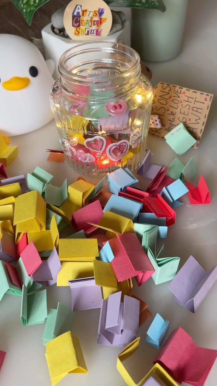 A glass jar with fairy lights surrounded by many small folded colorful paper notes.