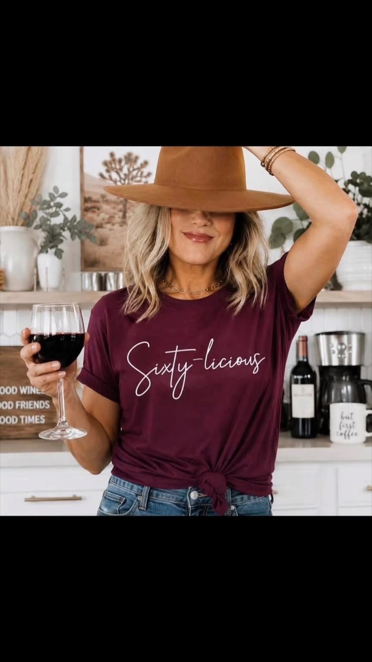 Woman in a maroon Sixty-licious t-shirt and hat holding a glass of red wine.