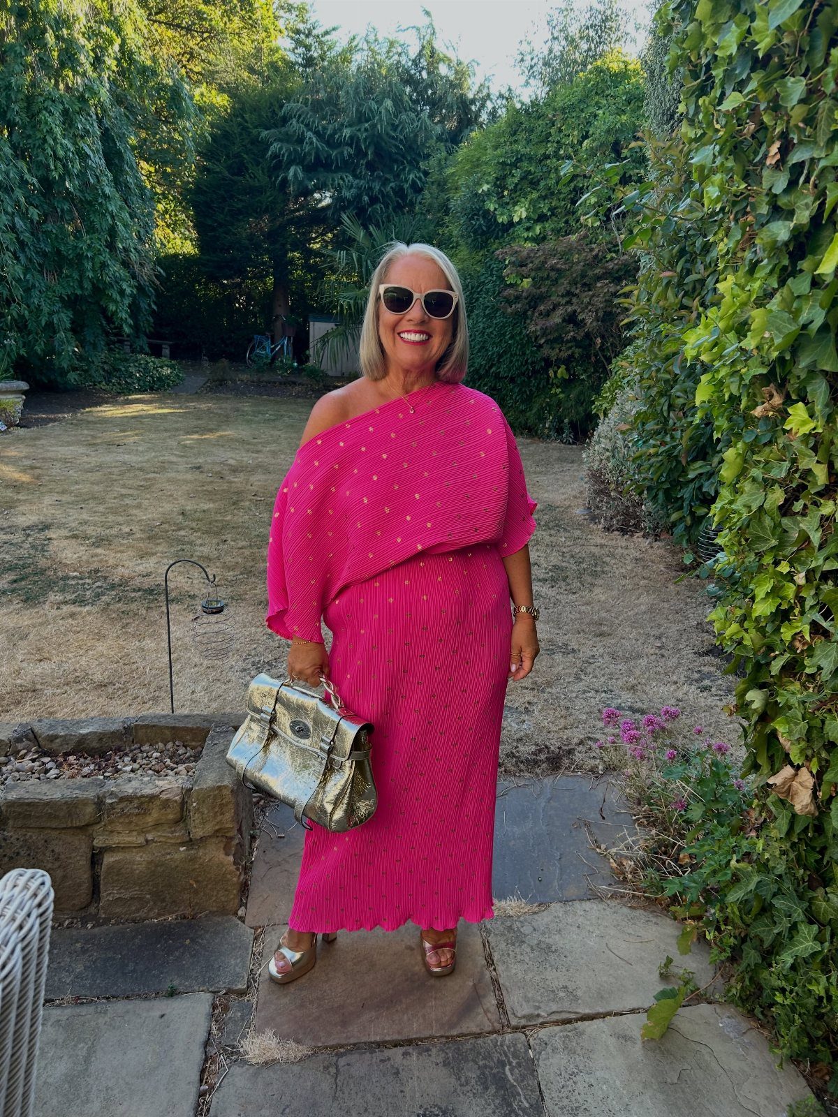A smiling woman in a pink one-shoulder maxi dress holding a silver handbag in a garden.