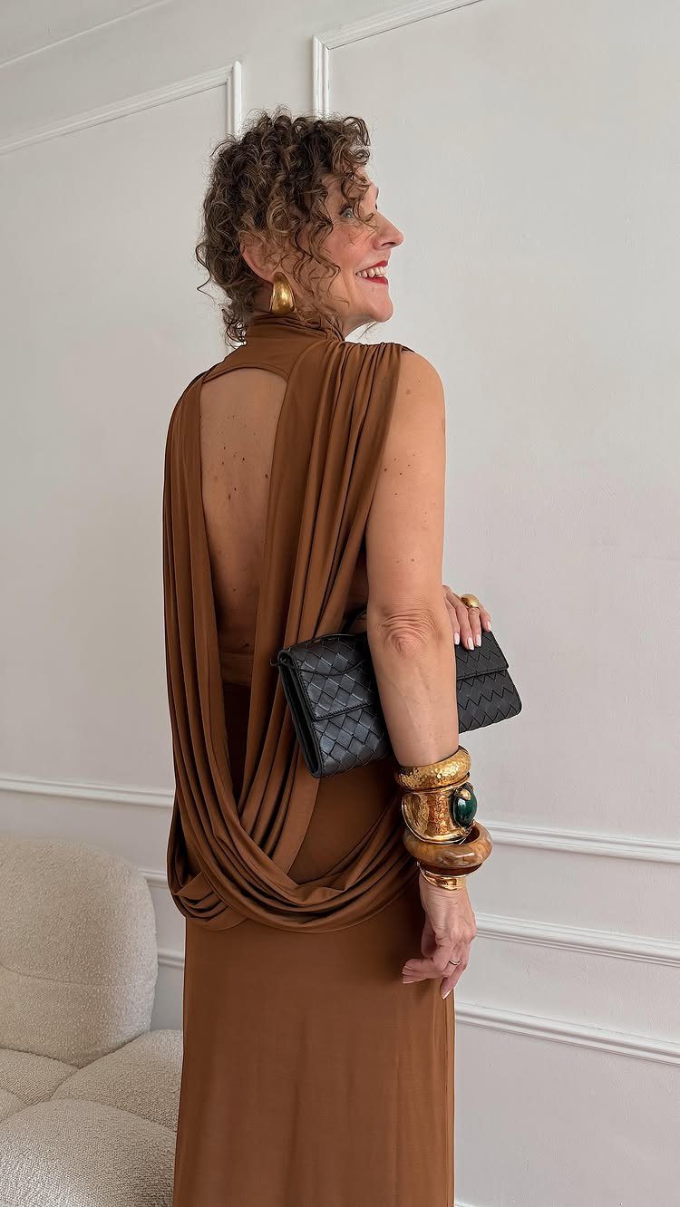 A woman in a brown draped maxi dress and gold jewelry standing against a white wall.