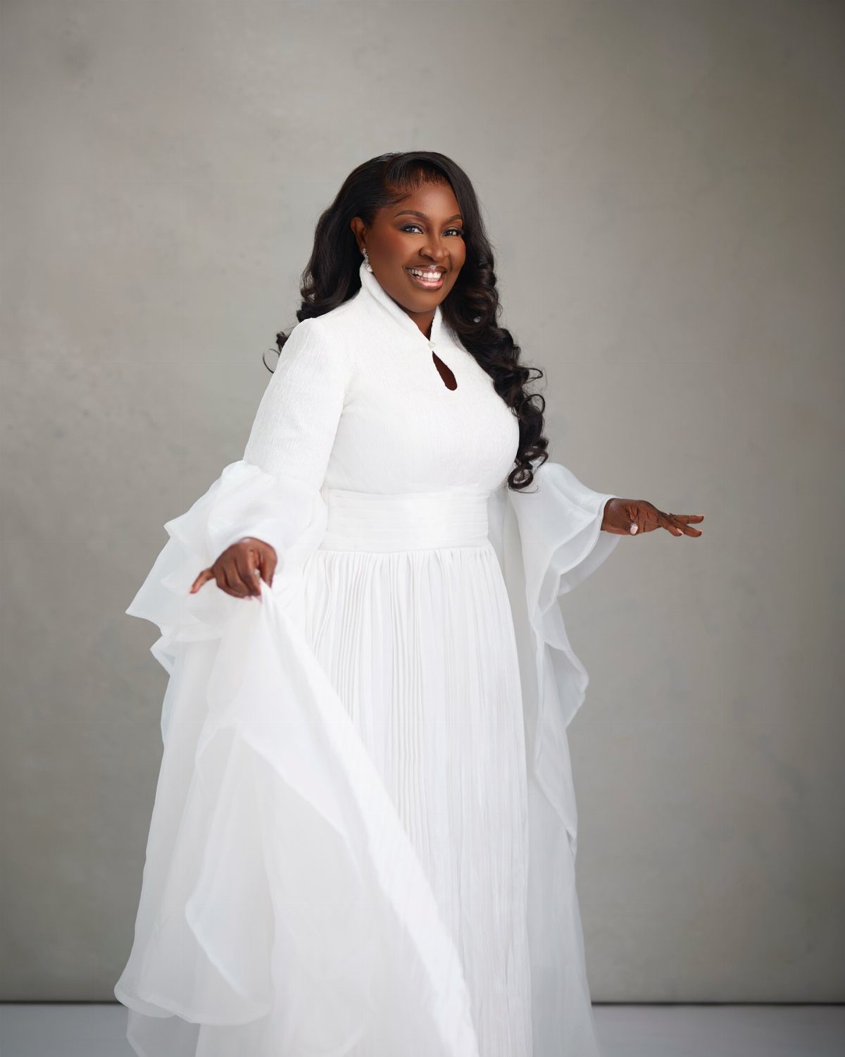 A smiling woman wearing an elegant, long-sleeved white formal gown with flowing ruffled sleeves.