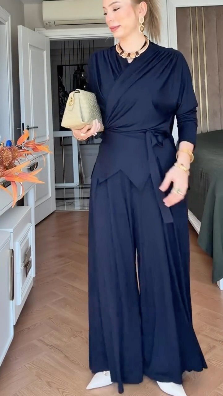A woman posing in a navy blue long-sleeved wrap jumpsuit and gold accessories.