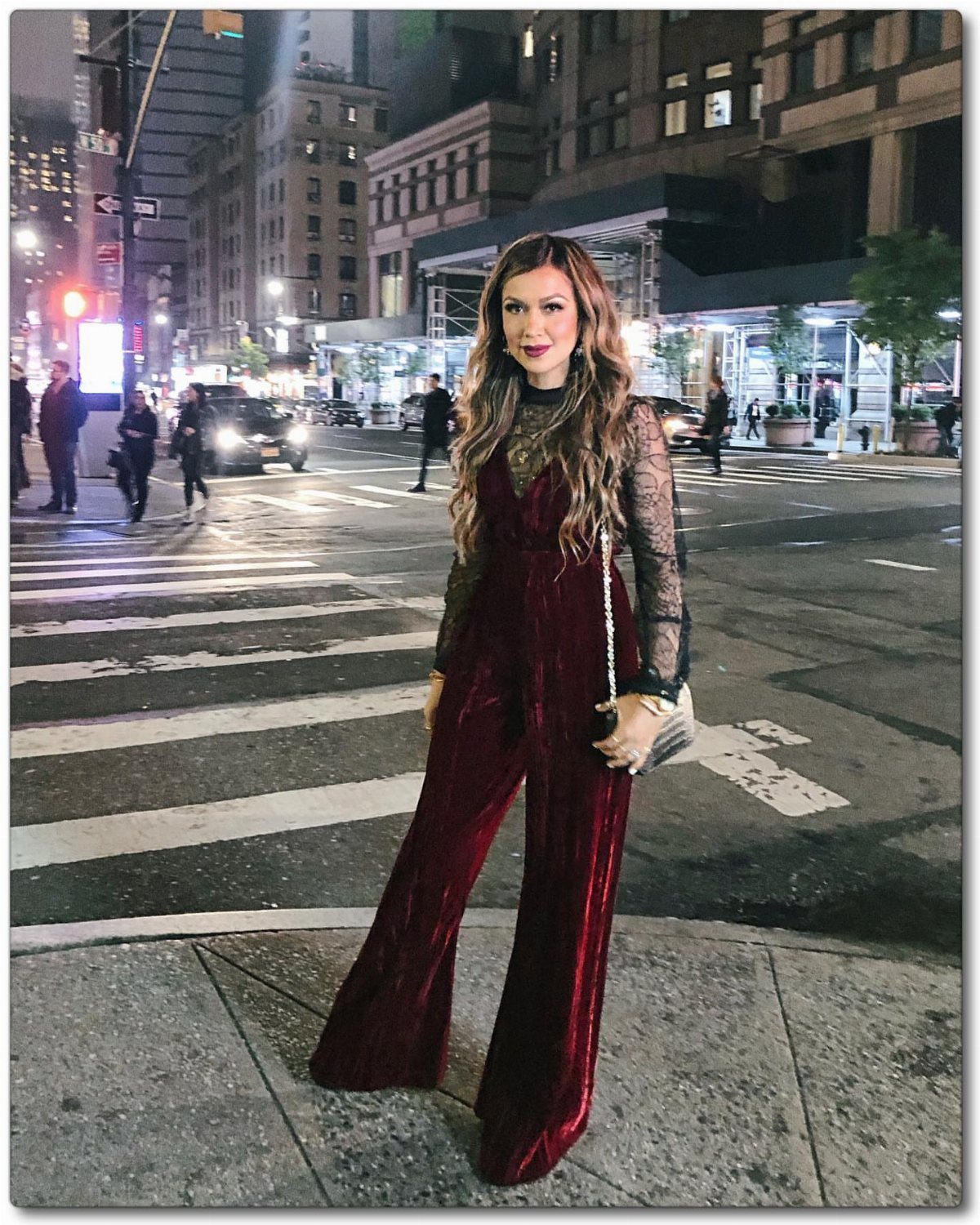 Woman in a dark red velvet jumpsuit with lace sleeves standing on a city street at night.