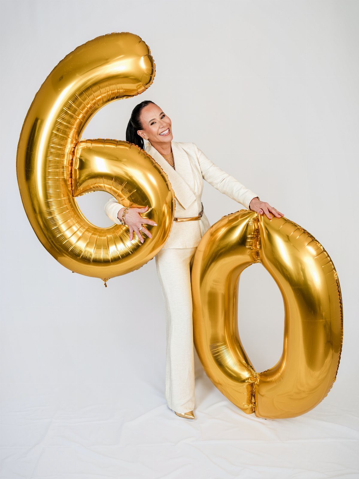 Smiling woman in a white blazer and trousers posing with large gold number sixty balloons.