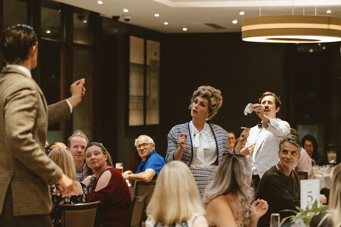 Actors performing a comedic scene for seated guests in a restaurant setting.