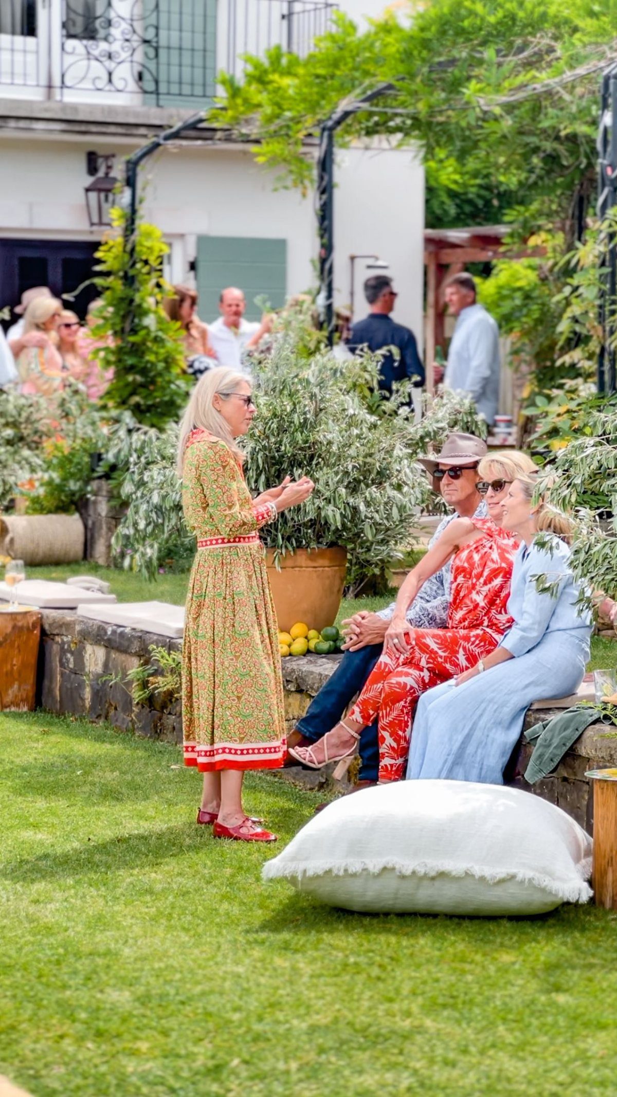 People socializing in a lush green garden setting with stone walls and potted plants.