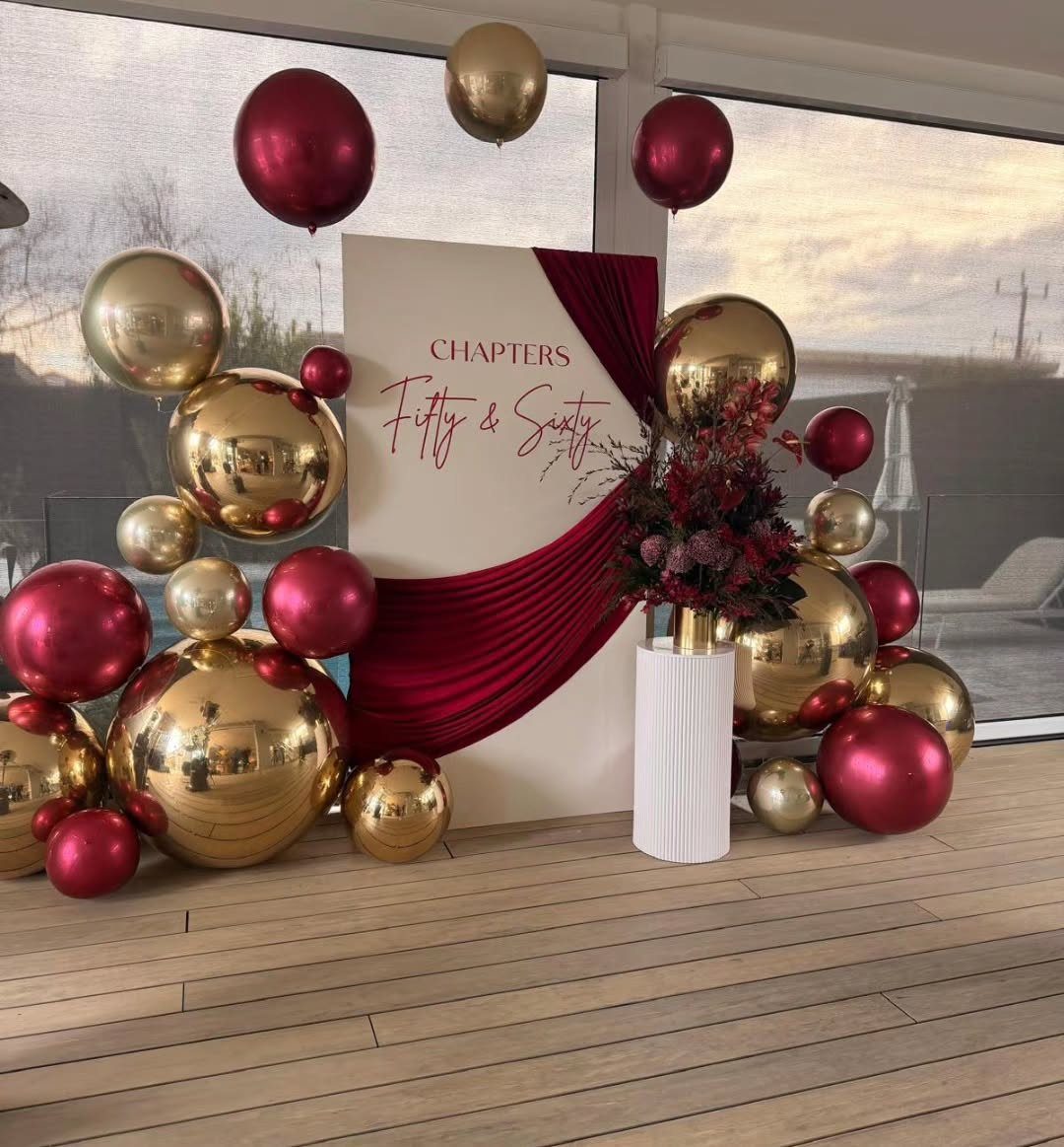 A decorative party backdrop with red and gold balloons surrounding a sign for a joint birthday.