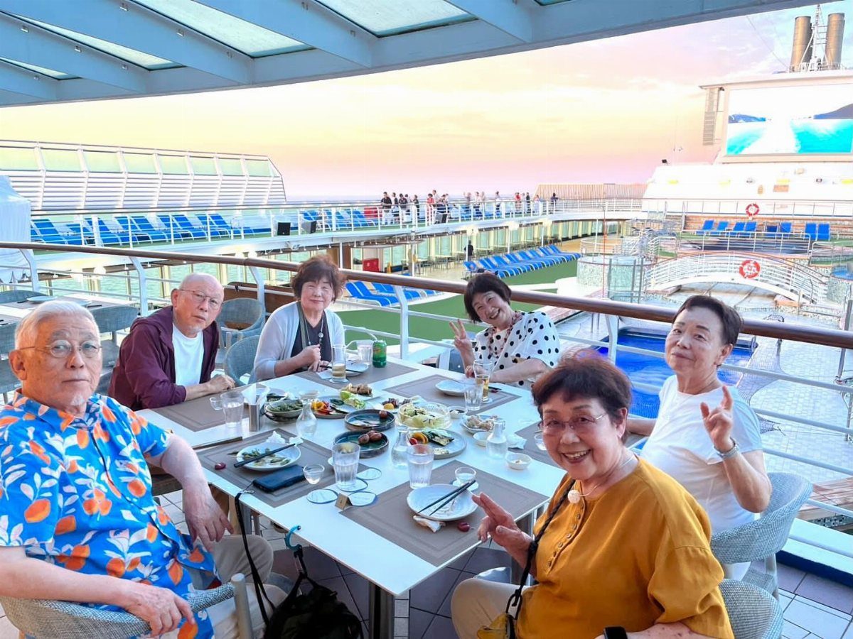 Elderly friends dining together on an outdoor deck of a large cruise ship at sunset.
