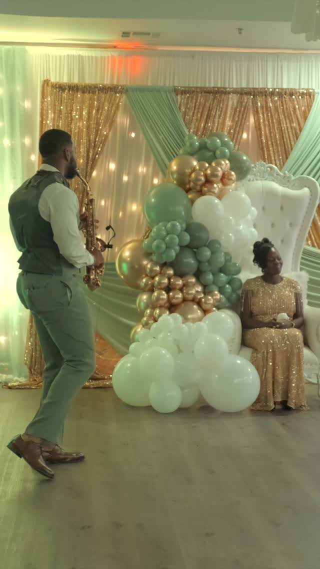 A man playing the saxophone for a woman seated in a white chair decorated with balloons.