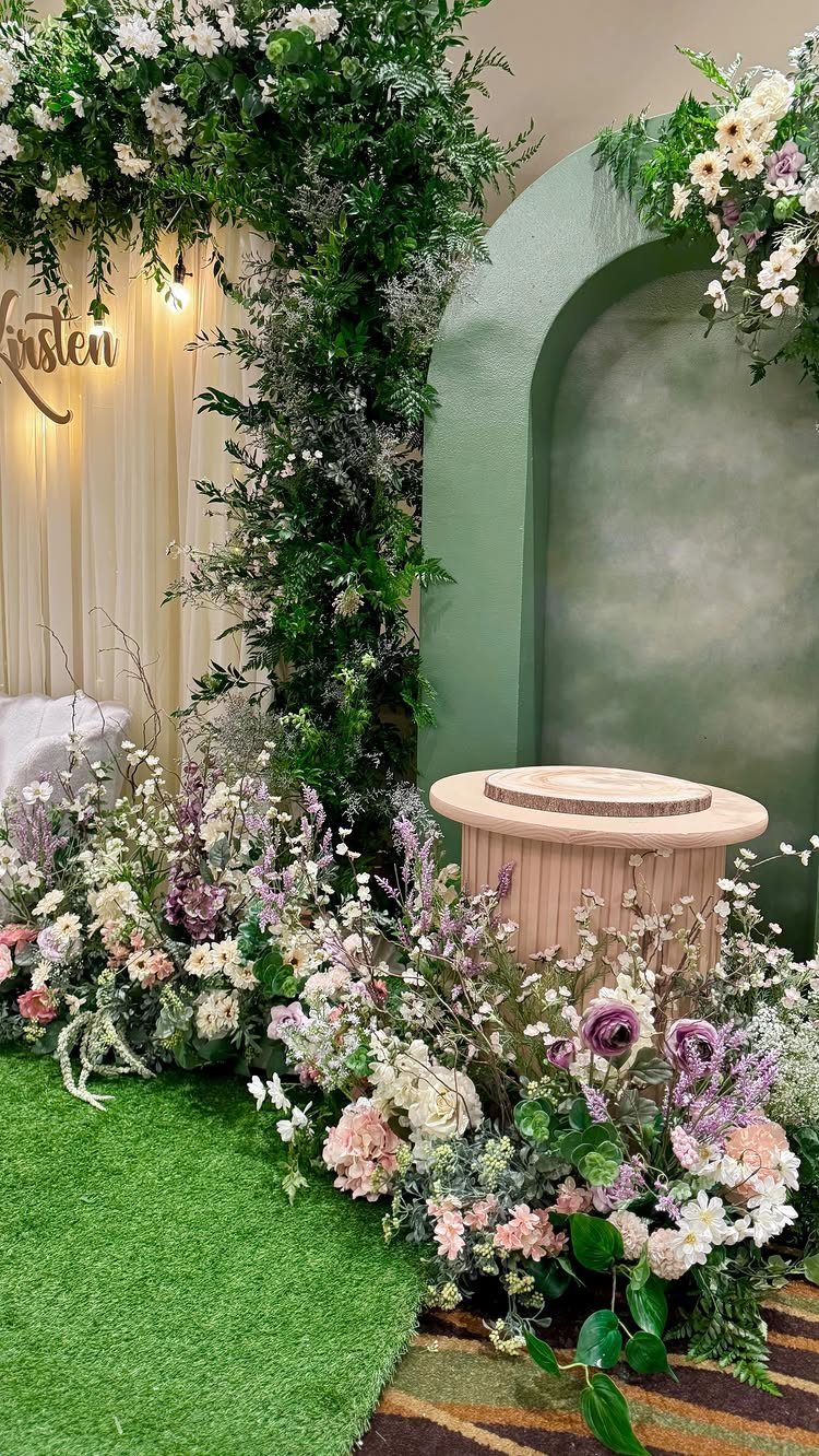 Floral arrangements and greenery decorate an event space with artificial grass and a pink pedestal.