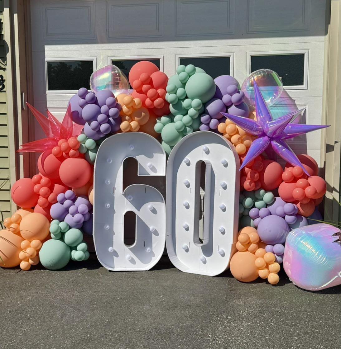 Large white marquee numbers sixty surrounded by a colorful balloon arch in front of a garage.