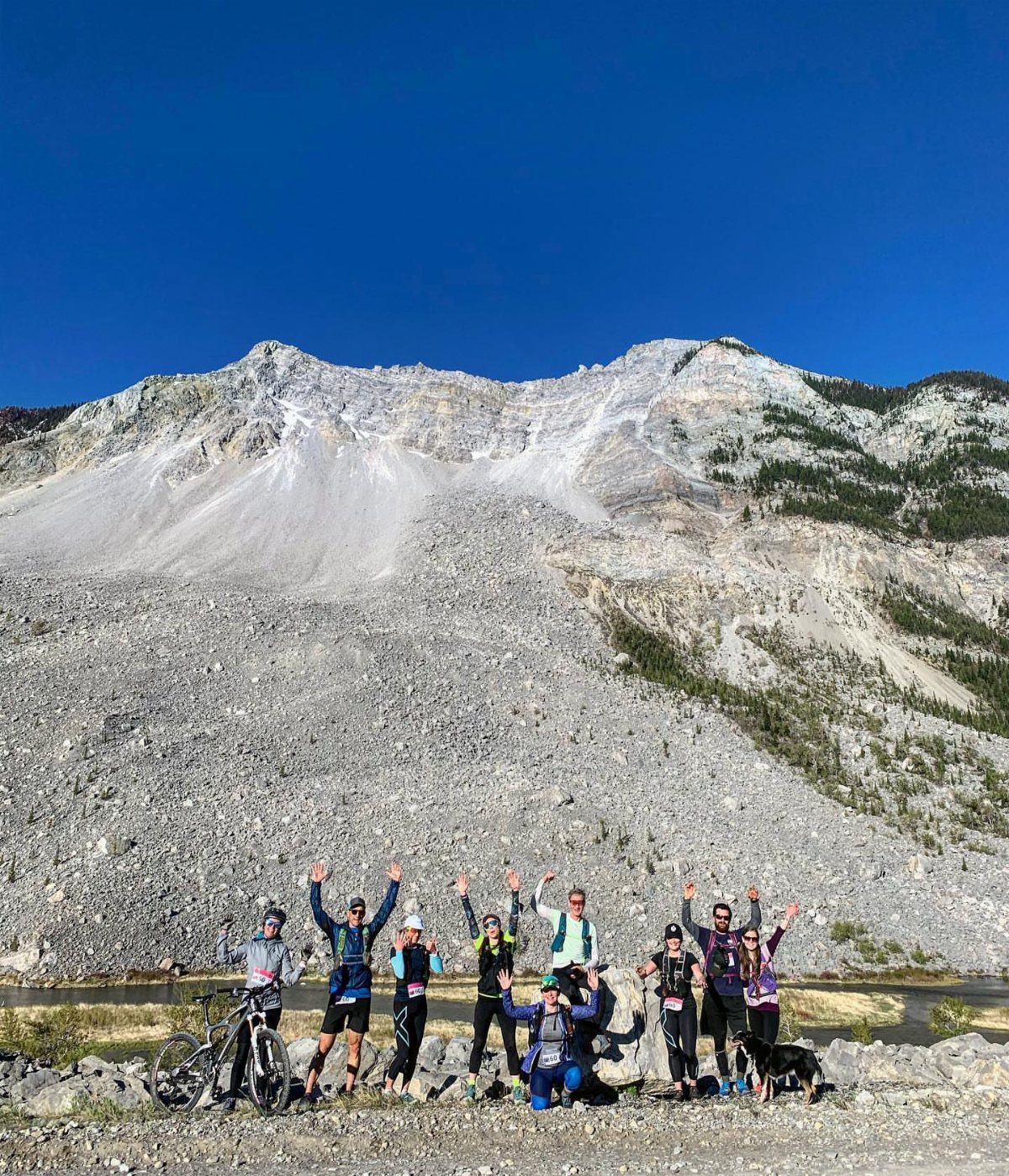 A group of hikers and cyclists posing with raised arms in front of a large rocky mountain.