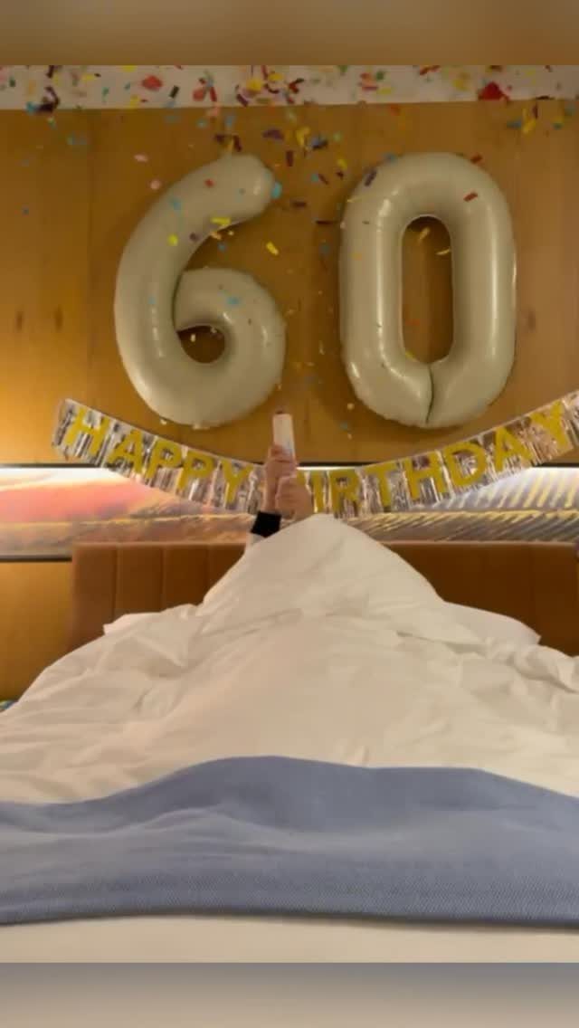 Large white number 60 balloons and a birthday banner hanging above a bed with confetti.
