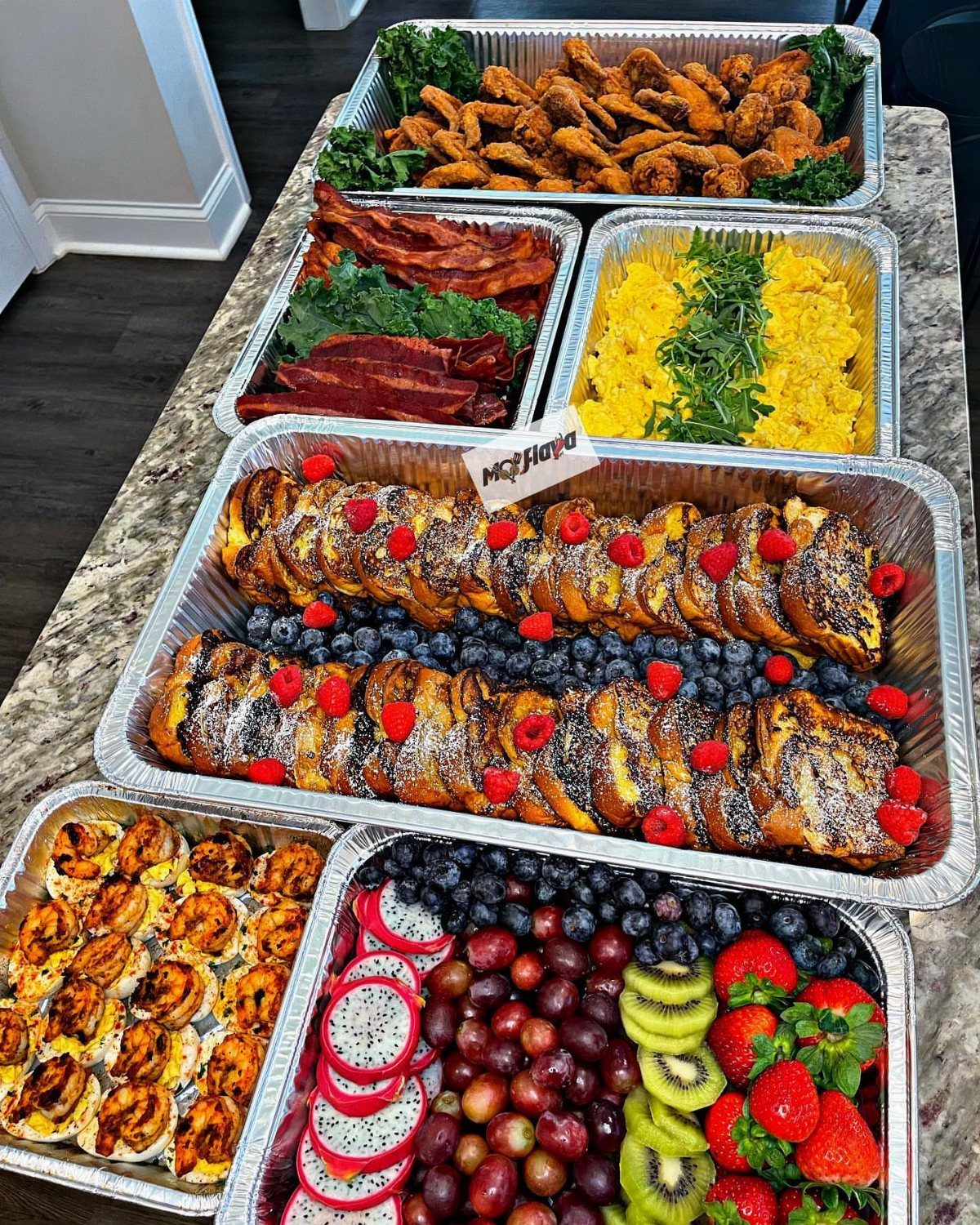 Various brunch food trays including French toast, fruit, eggs, and bacon on a granite counter.