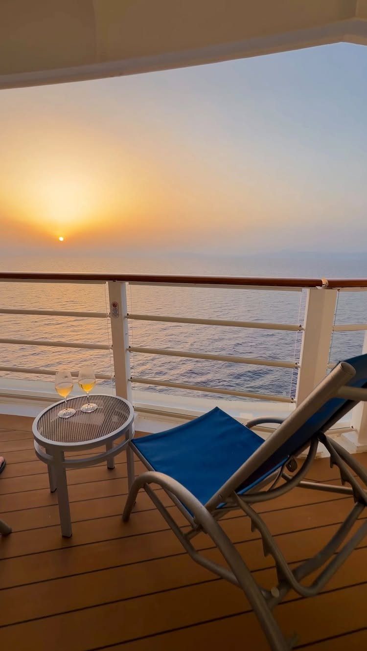 A blue lounge chair and drinks on a cruise ship balcony overlooking a sunset ocean.