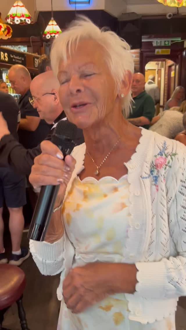 An older woman singing into a microphone during a social gathering indoors.