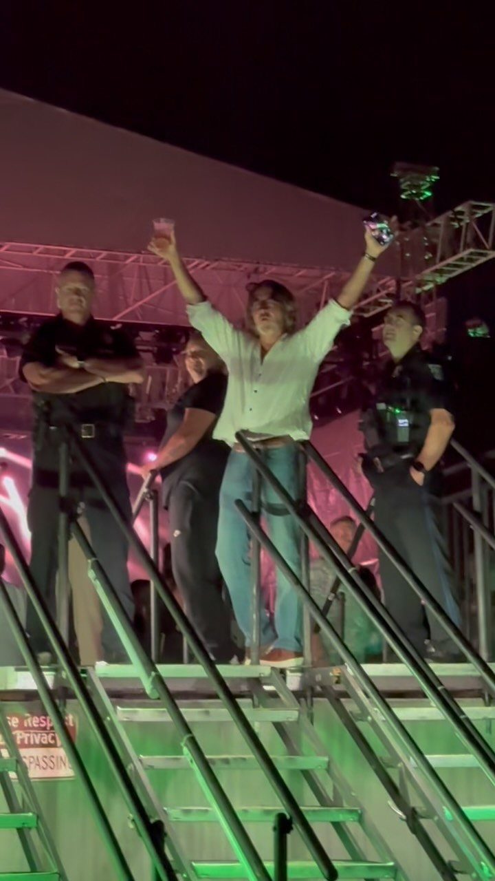 A male performer singing on a stage with colorful lights and security personnel present.