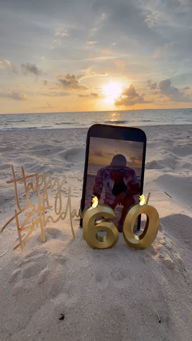 Gold number 60 candles and birthday sign on a beach at sunset.