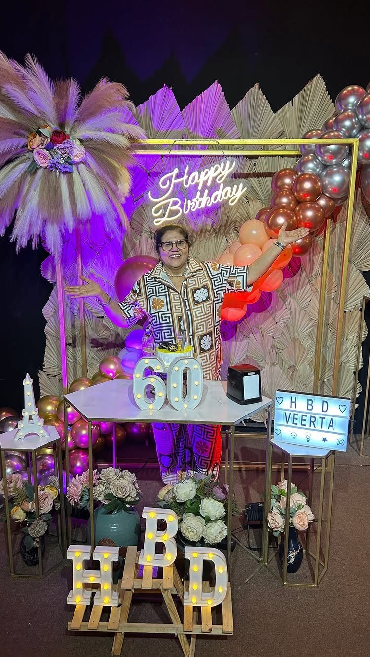 A woman smiling in front of a decorated 60th birthday backdrop with balloons and lights.