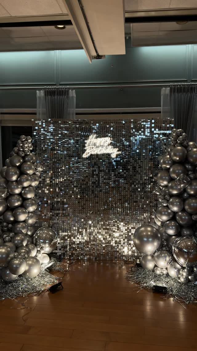 Silver sequin backdrop with large balloon arches and Happy Birthday text for a celebration.