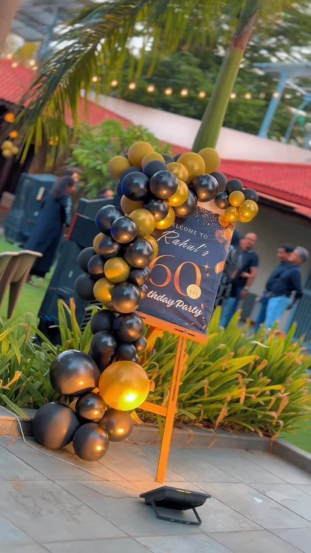Black and gold balloon garland decorating a welcome sign for a 60th birthday party outdoors.