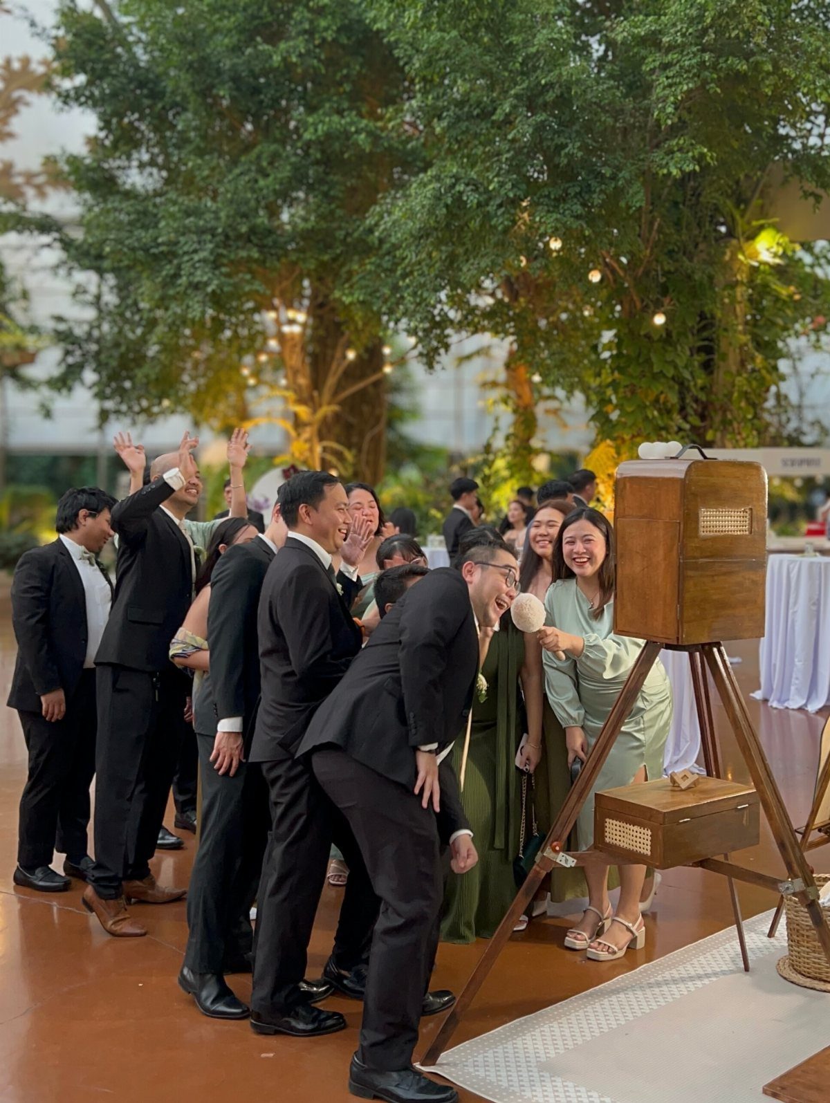 Group of guests leaning toward a vintage wooden audio guestbook on a tripod outdoors.