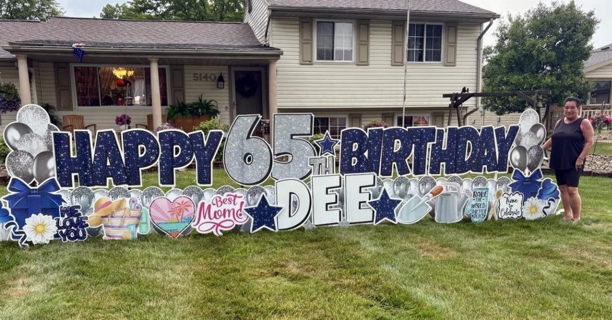 Large blue and silver glittery yard sign celebrating Dee's 65th birthday on a green lawn.