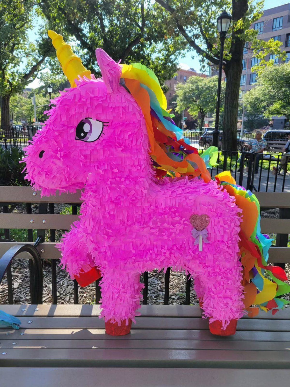 A bright pink unicorn piñata with a rainbow mane sitting on a park bench.