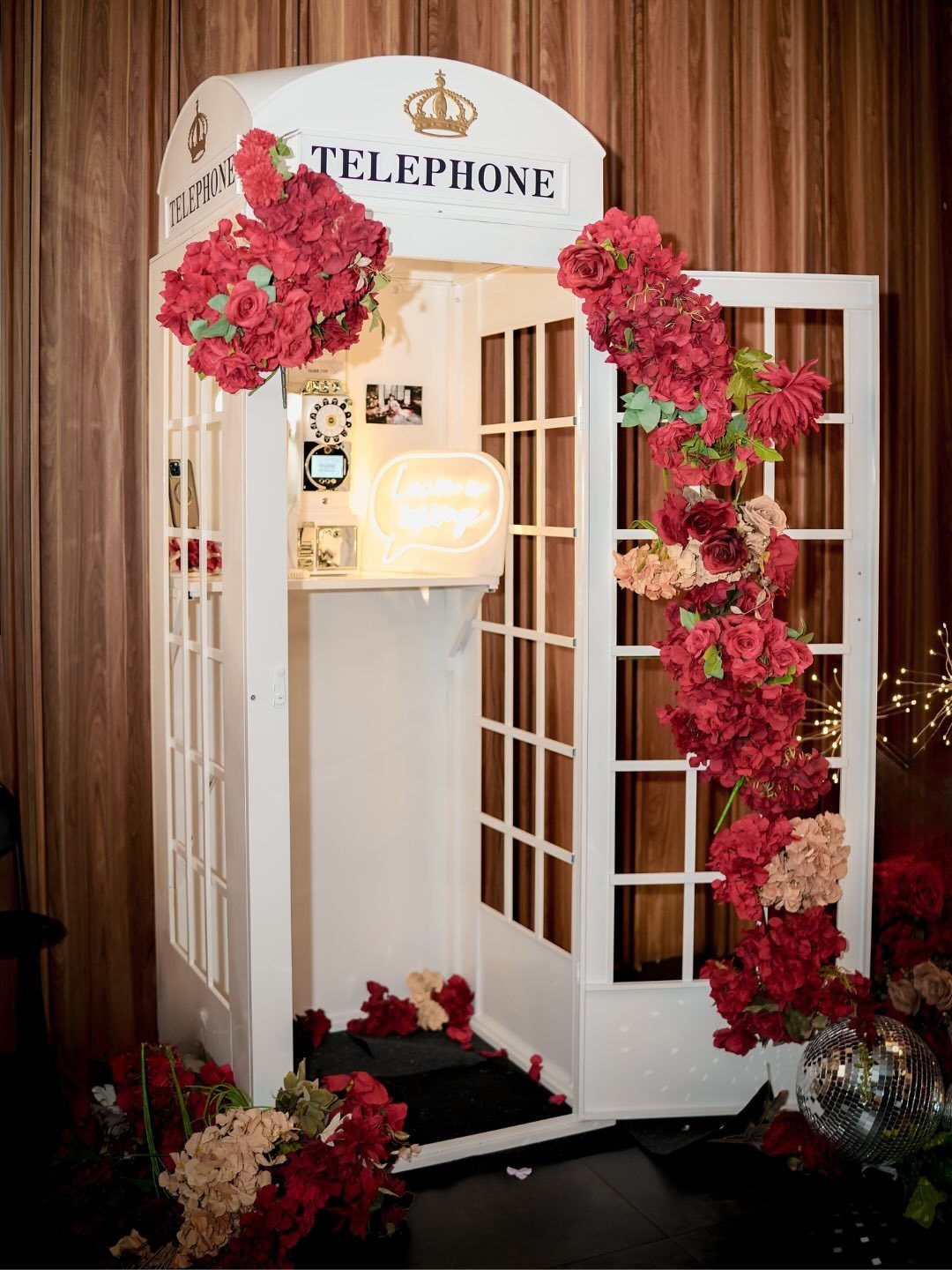 A white decorative telephone booth decorated with large red and pink floral arrangements.