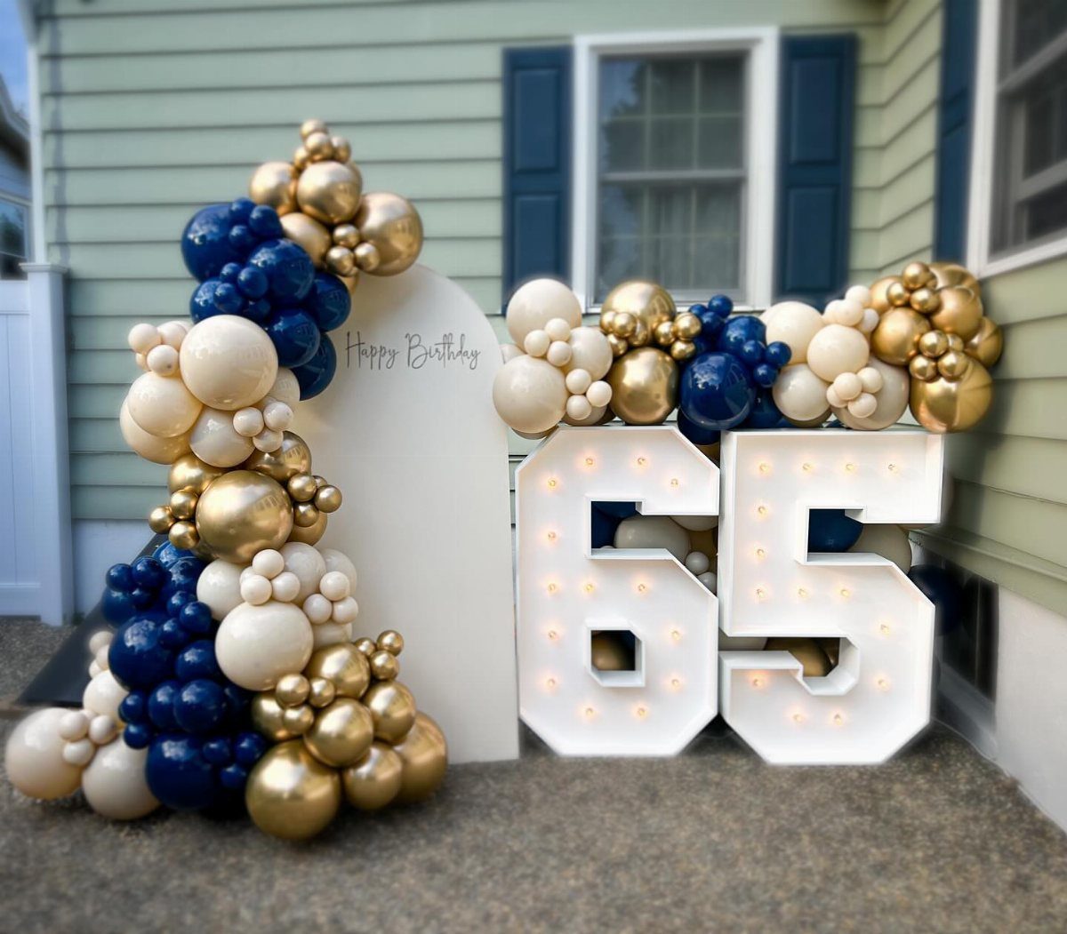 Large illuminated marquee numbers 65 surrounded by navy, gold, and white balloon garlands.