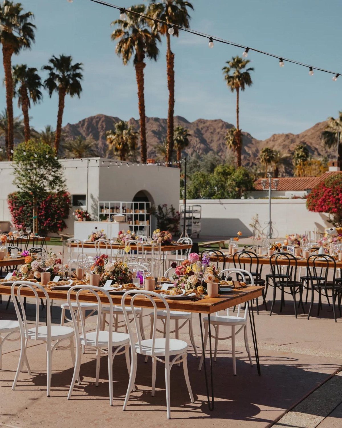 Long wooden dining tables with white chairs and floral arrangements set outdoors near palm trees.