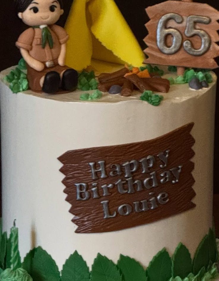 Decorated birthday cake with a scout figurine, yellow tent, campfire, and a 65 birthday sign.