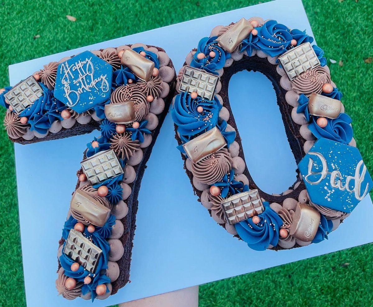 Chocolate number 70 cake decorated with blue frosting, chocolate bars, and gold candies on grass.