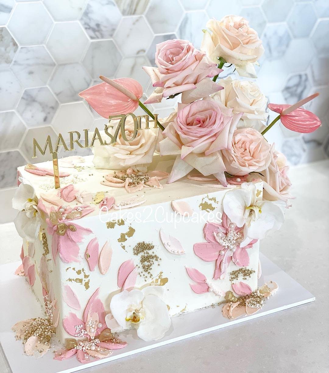 Square white cake decorated with pink buttercream flowers, gold leaf, and real roses.