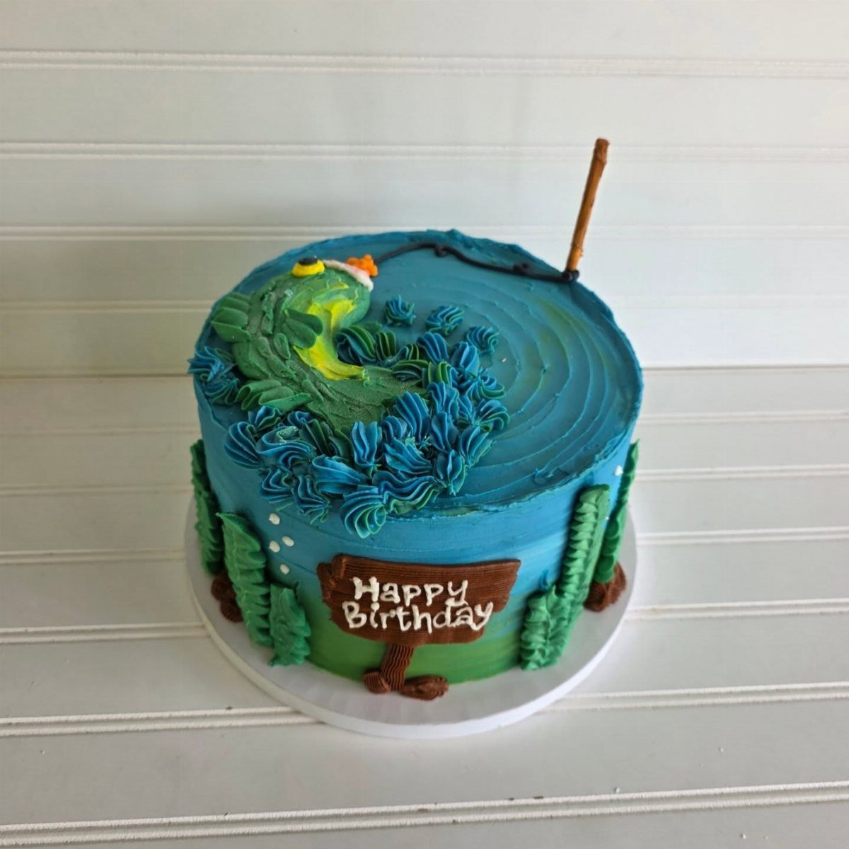 Blue frosted cake decorated with green waves, a small fish, and a fishing rod.