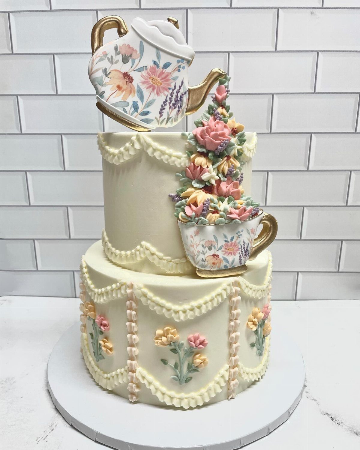Two-tier cream cake decorated with floral patterns, a large teapot on top, and a teacup overflowing with flowers.